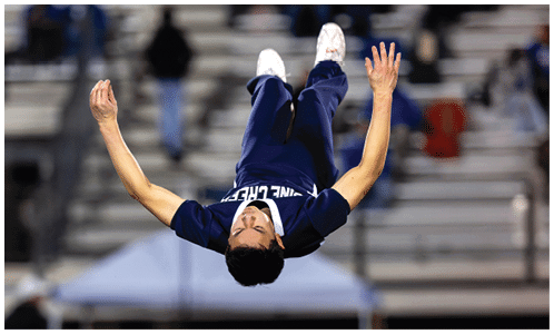A person in a sports uniform is captured mid-backflip on a field while another person in a similar uniform crouches in the background near bleachers. Description generated by AI