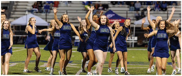 A group of people in blue cheerleading uniforms are performing a synchronized dance routine on a football field with spectators in the background. Description generated by AI