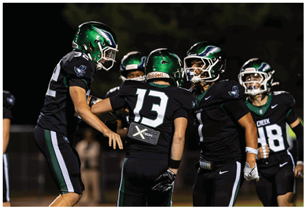 Four people wearing black football uniforms with green helmets are gathered on a field at night, appearing to celebrate or strategize. Description generated by AI