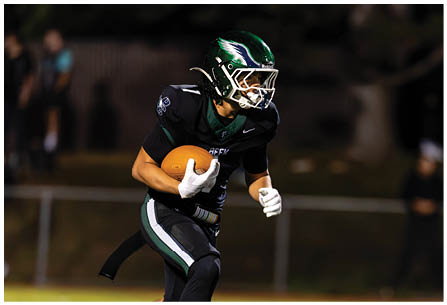 A person wearing a black and green football uniform is running on a grassy field while holding a football. Description generated by AI