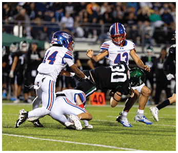 Three people in white and blue football uniforms tackle a person in a black football uniform on a grassy field during a game. Description generated by AI