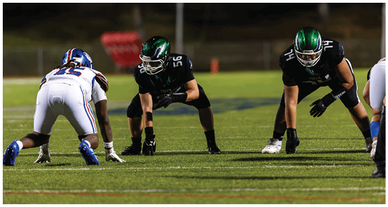 Three people in football uniforms are positioned on a field, appearing ready for a play during a game at night. Description generated by AI