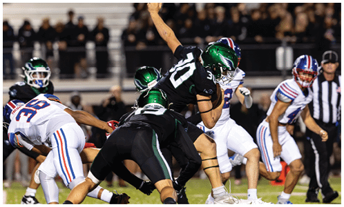 Players in black and white football uniforms with green helmets are engaged in a play on a grassy field with a referee and other players in the background. Description generated by AI