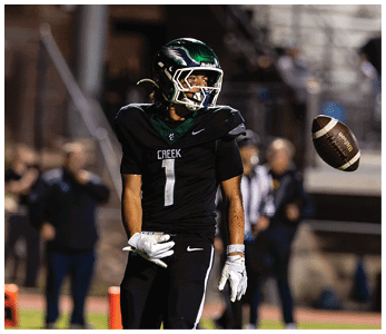 A person in a black football uniform with the word Creek and the number one is standing on a field while a football is mid-air nearby. Description generated by AI