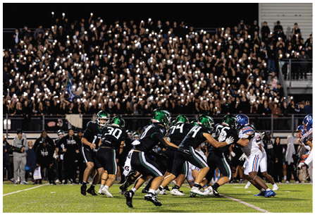A group of people in football uniforms are actively playing on a field with a large crowd in the background holding up lights. Description generated by AI