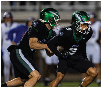 Two football players in black uniforms with green helmets are engaged in a play on the field, with one handing the ball to the other. Description generated by AI