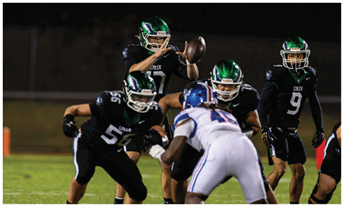 Several football players in black and green uniforms are positioned on a field with one player in white and blue uniform facing them during a game. Description generated by AI