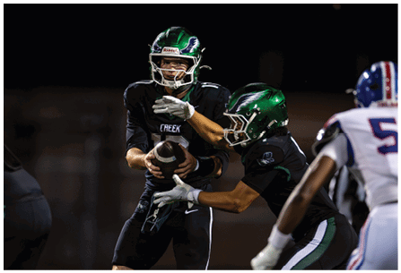 Two people in football uniforms with green helmets are engaged in a handoff during a nighttime game while a third player in a white uniform approaches. Description generated by AI