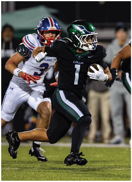 A person in a black football uniform with a green helmet is running with the ball while being chased by people in white uniforms on a football field during a night game. Description generated by AI