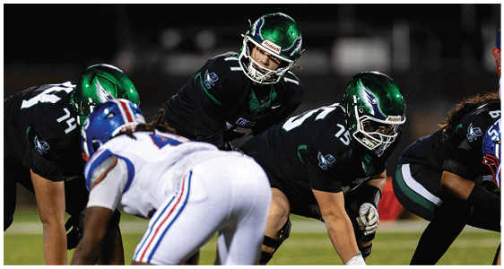 Four football players in helmets and uniforms are positioned on a field facing each other, preparing for a play during a game. Description generated by AI
