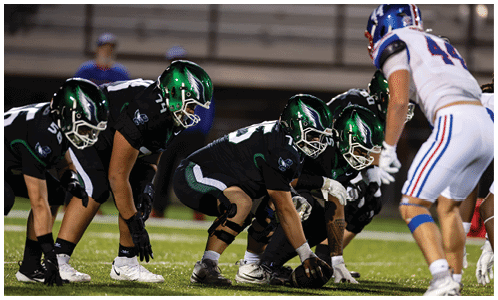 Several football players in black uniforms are lined up on the field facing a player in a white uniform during a game. Description generated by AI