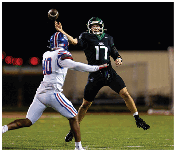 A person in a black sports uniform is throwing a football while another person in a white sports uniform attempts to block the pass on a grassy field at night. Description generated by AI