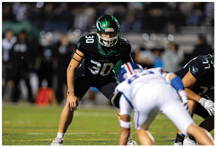 A person in a black football uniform with the number 30 is in a defensive stance facing two opposing players in white uniforms on a football field during a game. Description generated by AI