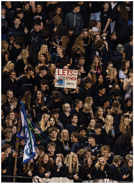 A large crowd of people wearing dark clothing is gathered closely together, with one person holding a sign that says “LETS GO SPENCER" and a flag visible in the lower left corner. Description generated by AI