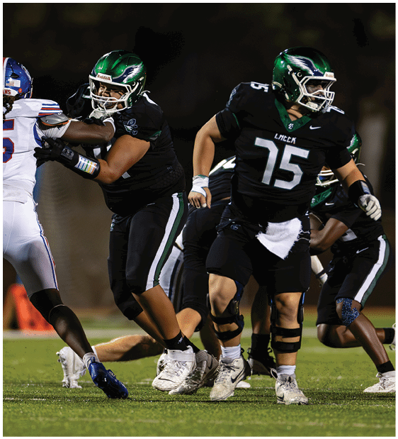 Two football players in black uniforms block a player in a white uniform during a game on a field at night. Description generated by AI