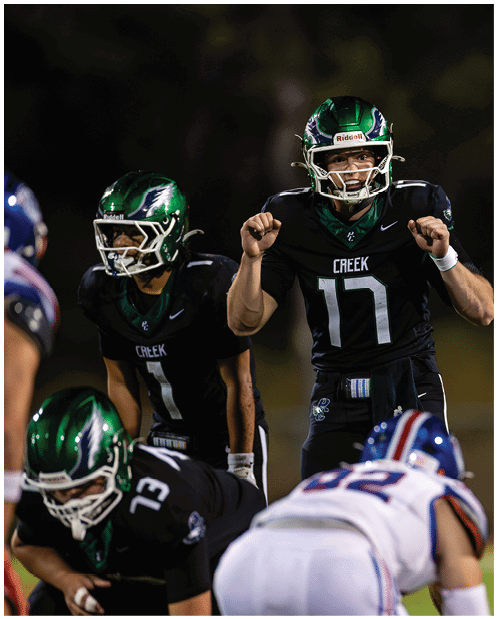 People wearing green and black football uniforms are positioned on a field, with one person in the center appearing to give instructions or signals. Description generated by AI