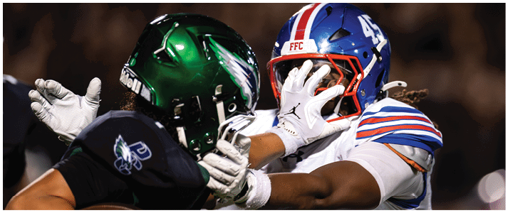 Two people in football uniforms and helmets are engaged in a close physical play during a game. Description generated by AI