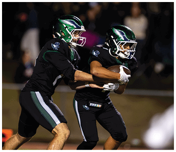 Two people wearing black football uniforms and green helmets are engaged in a handoff during a nighttime game. Description generated by AI