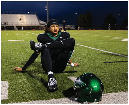 A person in a black football uniform is sitting on a field at night with one leg crossed over the other and a green helmet placed on the ground nearby. Description generated by AI
