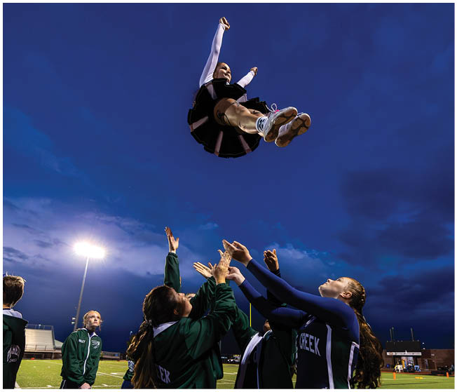 A person in a cheerleading outfit is being thrown high into the air by a group of people reaching up under a darkening sky with stadium lights in the background. Description generated by AI