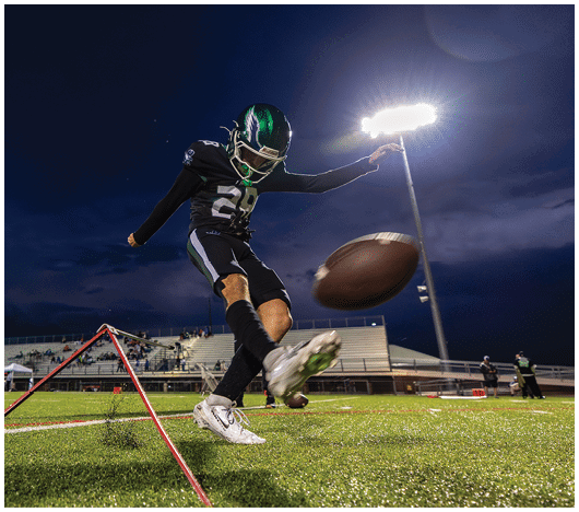 A person wearing a sports uniform is kicking a football on a grassy field under a dark evening sky with stadium lights and empty bleachers in the background. Description generated by AI
