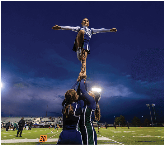 A person in a cheerleading uniform is being lifted into the air with one leg raised and the word “HOOPS" written vertically on their raised leg against a clear blue sky. Description generated by AI