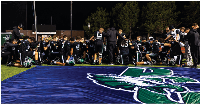 A group of people wearing black sports uniforms are gathered closely together on a field at night near a large blue banner with a green and white logo. Description generated by AI