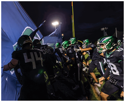 A group of football players wearing green helmets and dark uniforms run energetically onto the field through a large blue flag at night under stadium lights. Description generated by AI