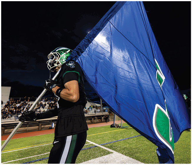 A person in a sports uniform and helmet is holding and waving a large blue flag on a football field at night with spectators in the background. Description generated by AI