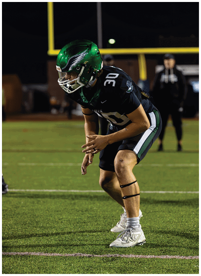 A person wearing a green football helmet and black jersey with the number 30 is leaning forward in a ready position on a football field. Description generated by AI