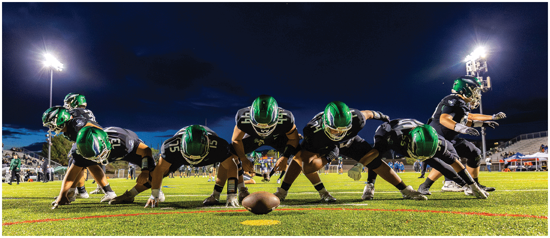 A group of football players in green helmets and black uniforms are lined up on a field at night, preparing for a play. Description generated by AI