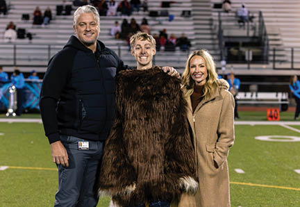 Three people stand on a football field with one person in the center wearing a large furry costume with yellow bird-like feet while the other two wear casual clothing and smile. Description generated by AI