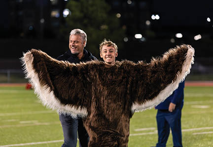 A person wearing a large furry costume with outstretched arms stands on a sports field at night while two other people stand nearby smiling. Description generated by AI