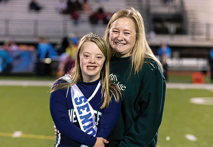 Two smiling people stand closely together on a sports field, one wearing a dark sweatshirt and the other in a cheerleading uniform with a sash and tiara. Description generated by AI