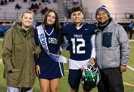 Four people stand together on a sports field with one person wearing a football uniform holding a helmet and another person wearing a cheerleading outfit with a sash. Description generated by AI