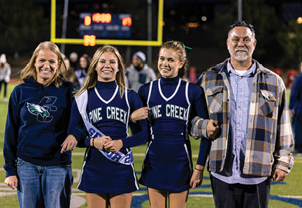 Two adults and two people in cheerleading uniforms stand linked arm in arm on a sports field at night with a scoreboard and goalpost in the background. Description generated by AI