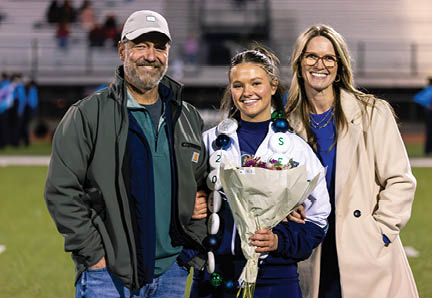 Three people stand closely together on a sports field with one person in the center holding a bouquet of flowers and wearing a sports jacket and a celebratory lei. Description generated by AI