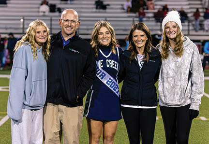 Five people stand closely together on a sports field at night, smiling, with one person wearing a cheerleading uniform and others dressed in casual and warm clothing. Description generated by AI