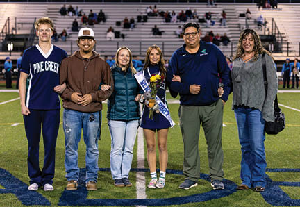 A group of six people standing on a sports field with bleachers in the background, with one person in the center wearing a sash and holding flowers and a stuffed animal. Description generated by AI
