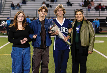 Four people stand linked arm-in-arm on a sports field with bleachers in the background, one person wearing a sports jersey and holding a bouquet of flowers. Description generated by AI