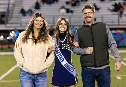 Three people stand together smiling on a sports field with one person in the center wearing a cheerleading uniform and a sash that reads “Senior." Description generated by AI