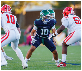 Three football players in action on a field with two wearing white uniforms and red helmets and one wearing a dark blue uniform and green helmet. Description generated by AI