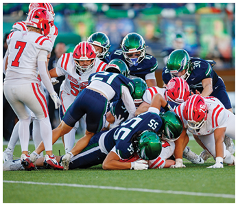 A group of people wearing football uniforms in red and white and others in dark blue and green are engaged in a close physical play on a grassy field. Description generated by AI