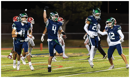 Four people in blue football uniforms celebrating on a field while others in red uniforms stand in the background. Description generated by AI