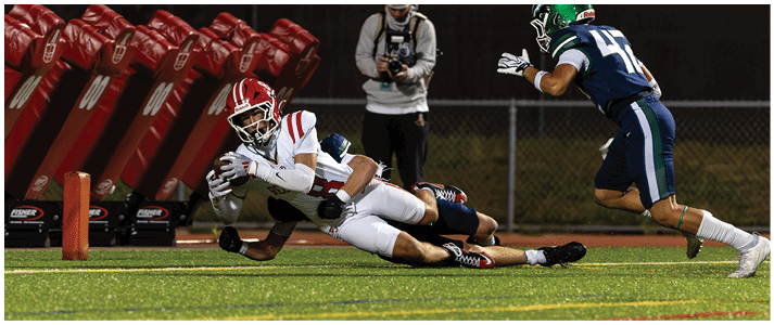 A person in a white football uniform is diving forward with the ball while another person in a dark uniform tackles them on a football field with a photographer in the background. Description generated by AI
