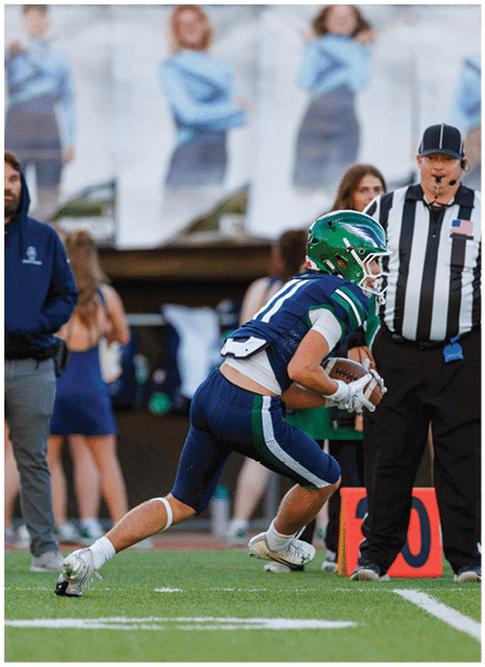 A person in a green helmet and blue sports uniform is running with a football on a field while a referee in a striped shirt watches nearby. Description generated by AI