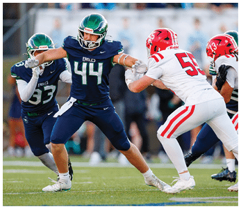 Two people in dark blue football uniforms with green helmets are blocking two people in white football uniforms with red helmets on a grassy field during a game. Description generated by AI