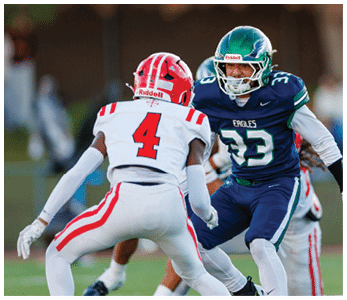 Two people in football uniforms are engaged in a play on a field, with one wearing a red and white uniform and the other in a blue and white uniform labeled Eagles. Description generated by AI