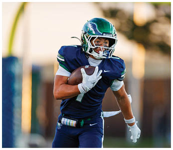 A person wearing a blue football uniform and helmet is running while holding a football on a field with a goalpost in the background. Description generated by AI