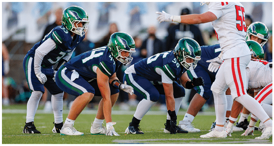 Four people in green and blue football uniforms are crouched in a ready position facing three people in white and red football uniforms on a grassy field. Description generated by AI
